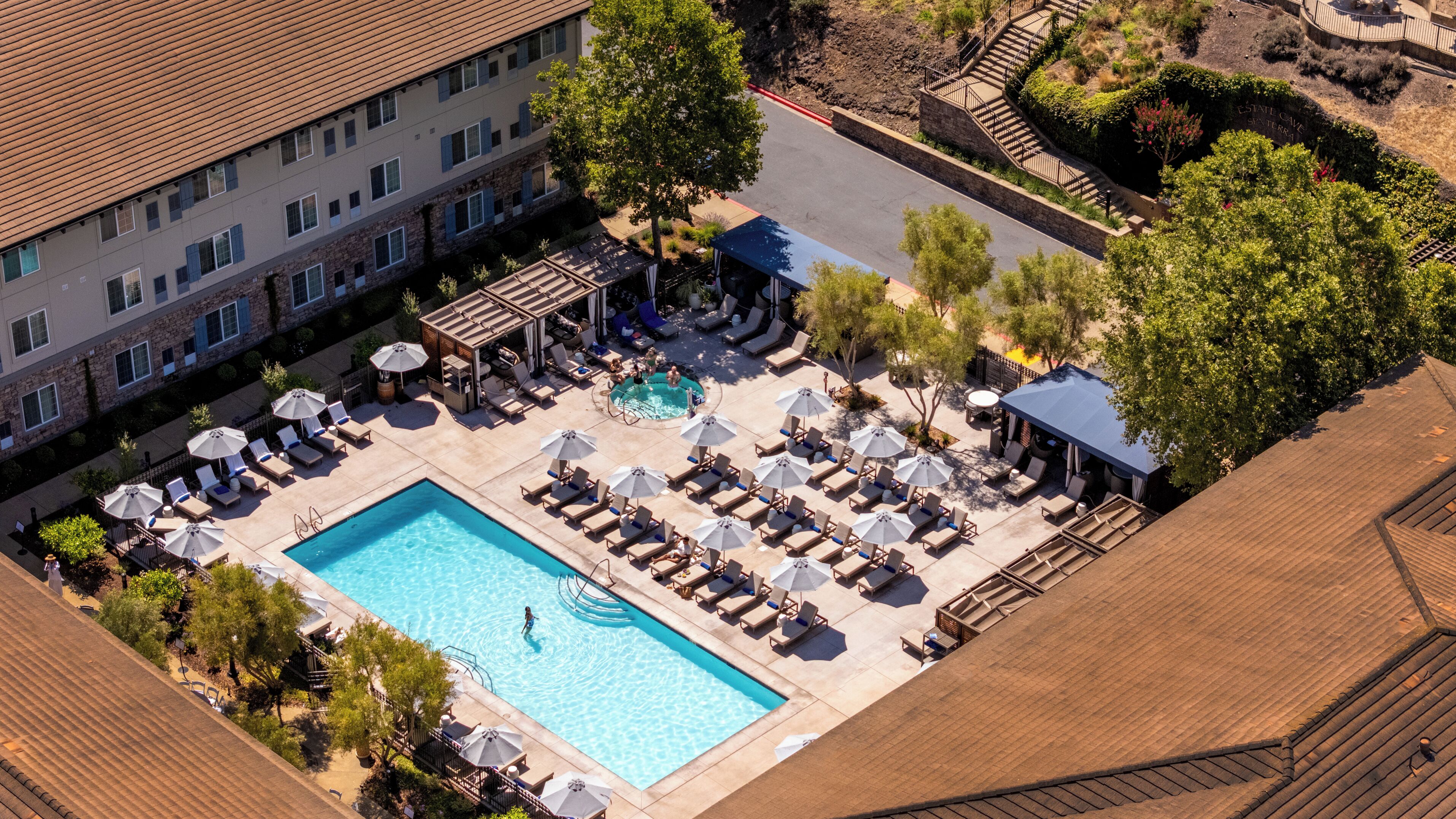 Vista aérea de un hotel con piscina rodeada de sillas de descanso y sombrillas, un área de estar techada y un jardín ajardinado.