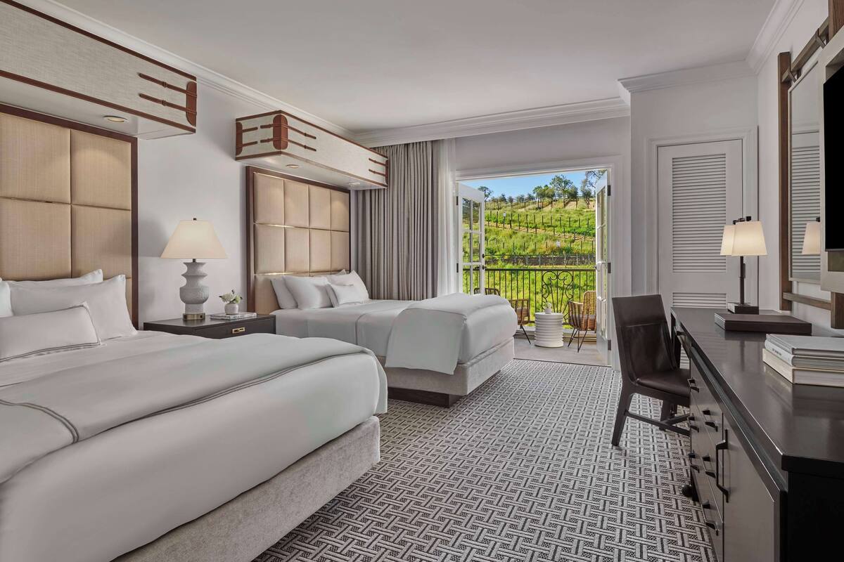 A hotel room with two beds, a desk, a chair, and a view of a vineyard through a large window.
