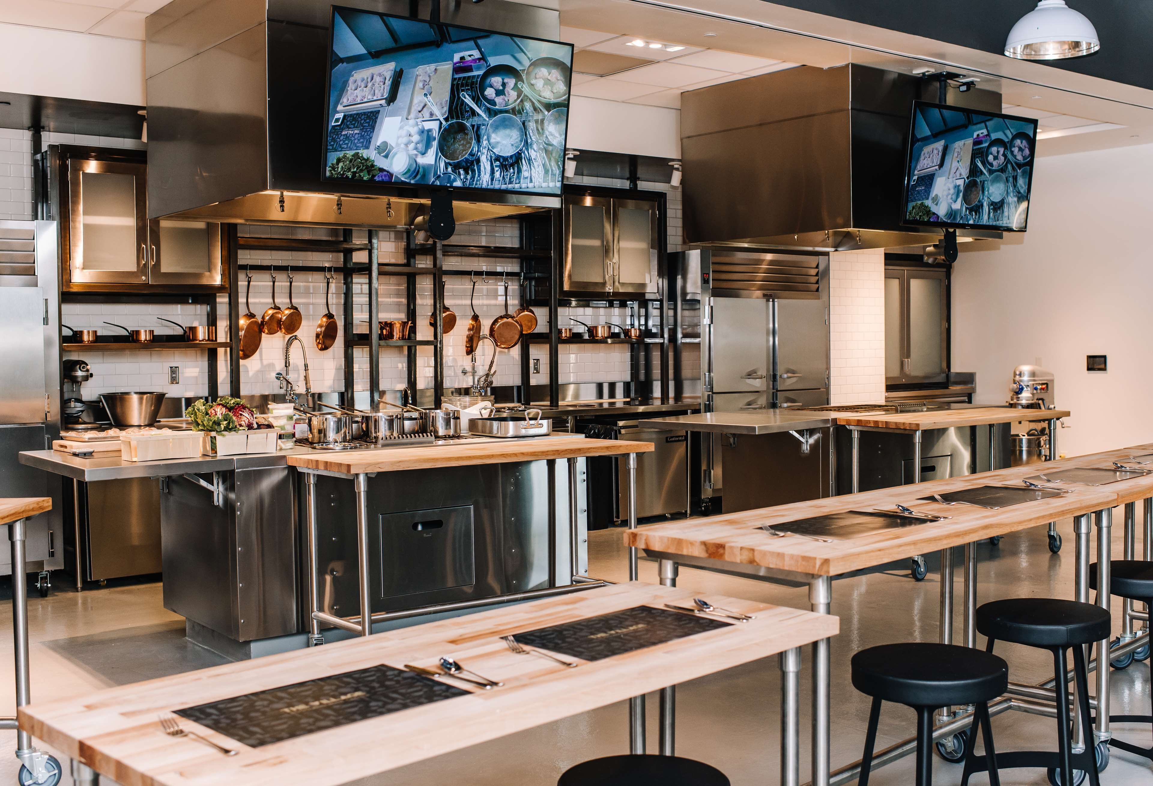 A modern kitchen with stainless steel appliances, wooden countertops, and two TVs mounted on the wall.