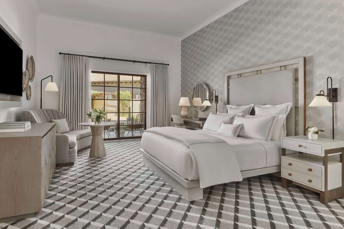 A modern hotel room with a large bed, a sofa, a desk, and a view of the outdoors.