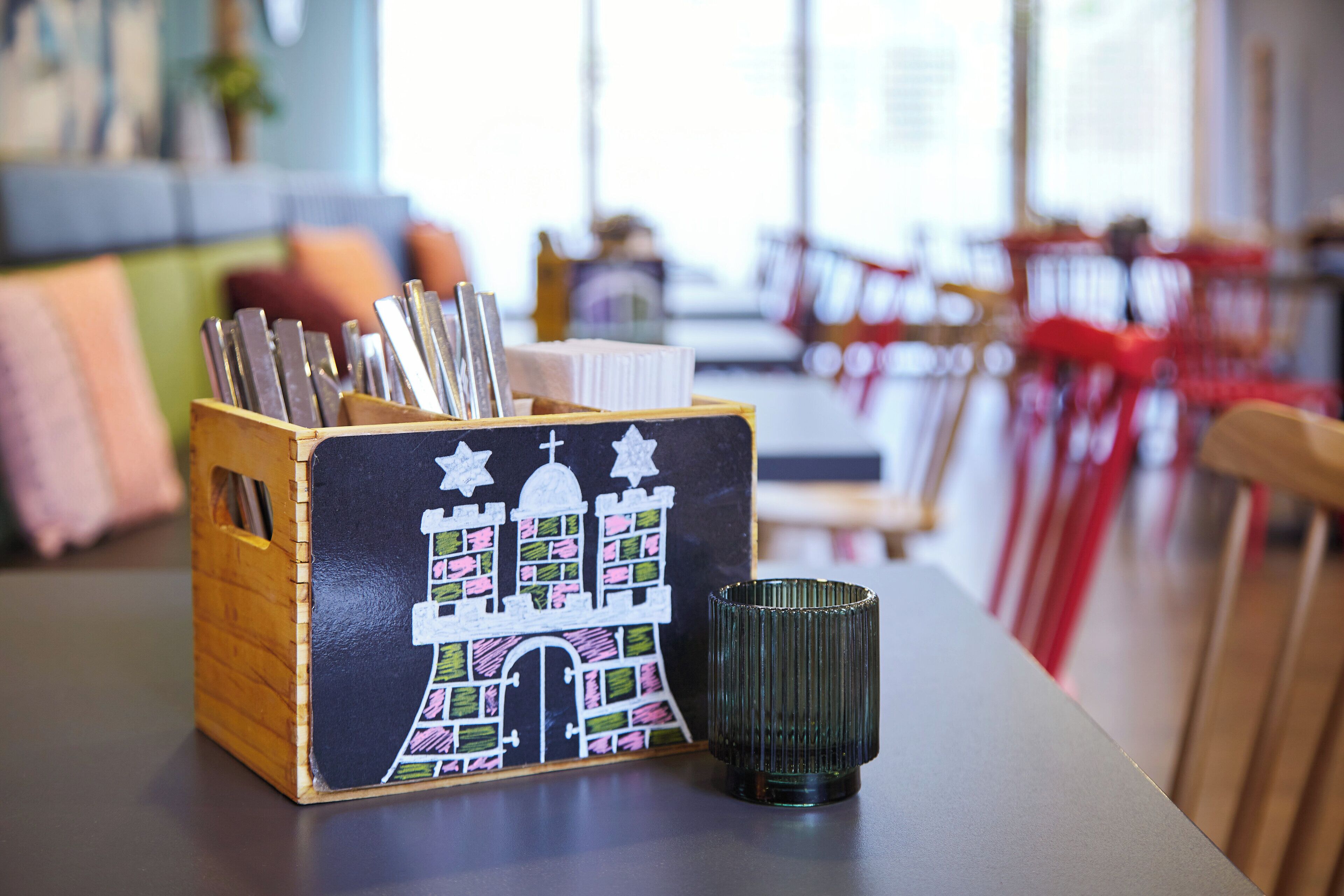 A wooden box with pens and pencils, a black mug, and a colorful building illustration on a table in a cafe setting.