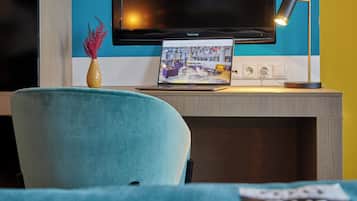 A modern room with a teal chair, a laptop on a desk, a vase with red flowers, and a flat-screen TV mounted on the wall.