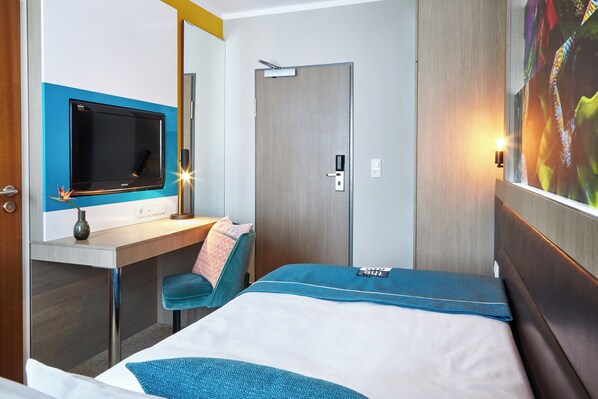 A hotel room with a bed, a desk with a flat-screen TV, a chair, and a door.
