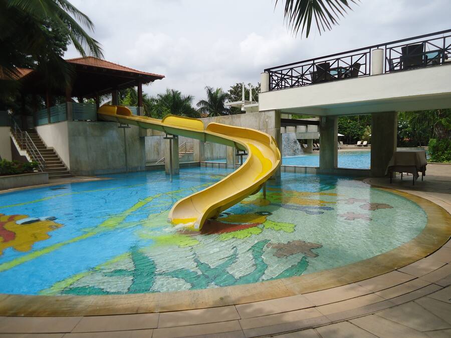 A swimming pool with a yellow slide, a covered area with stairs, and a view of a water fountain.