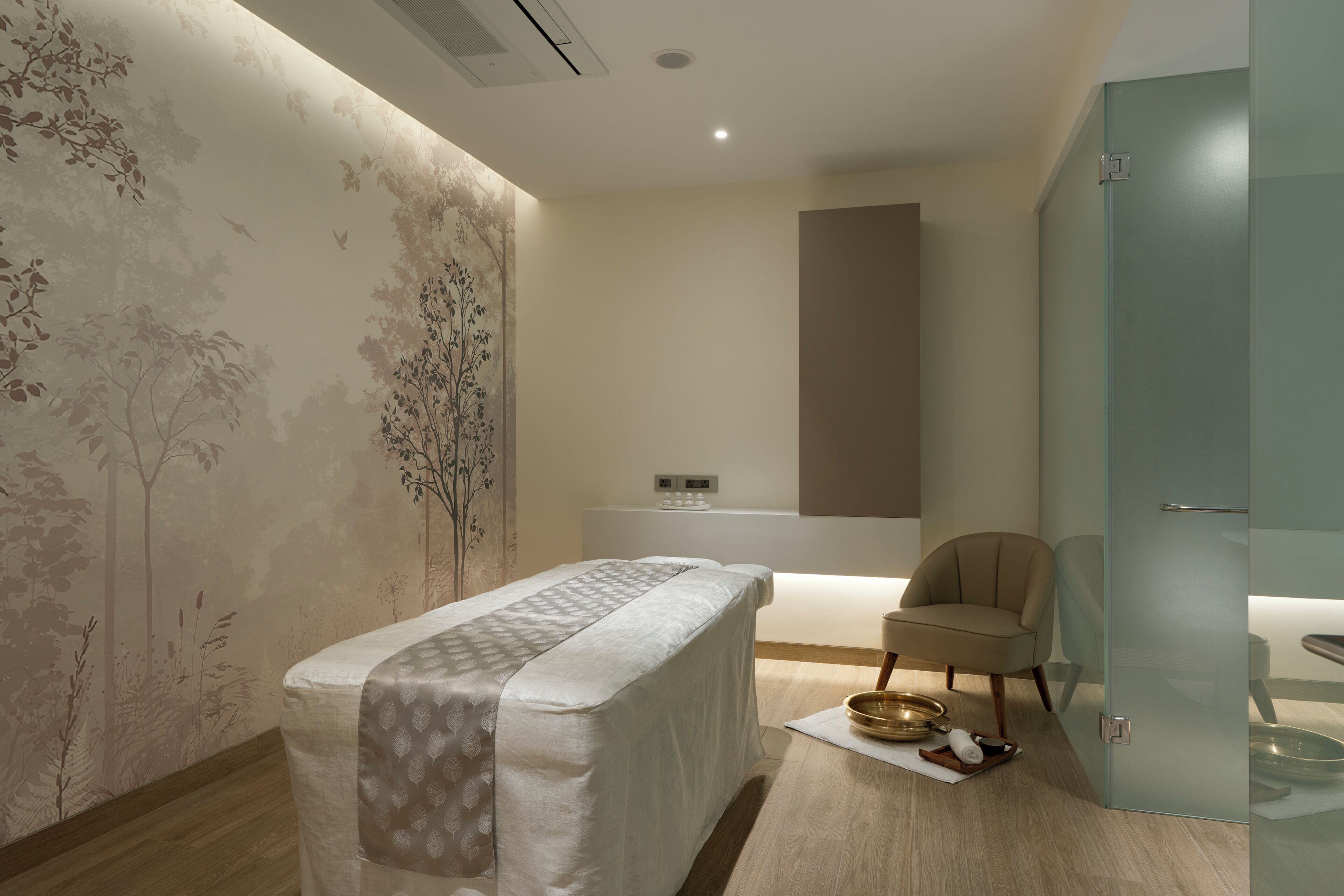 Ein Raum mit Massagetisch, einem Sessel, einem kleinen Tisch mit Schale und Handtuch sowie einer Wand mit Baumdekor.