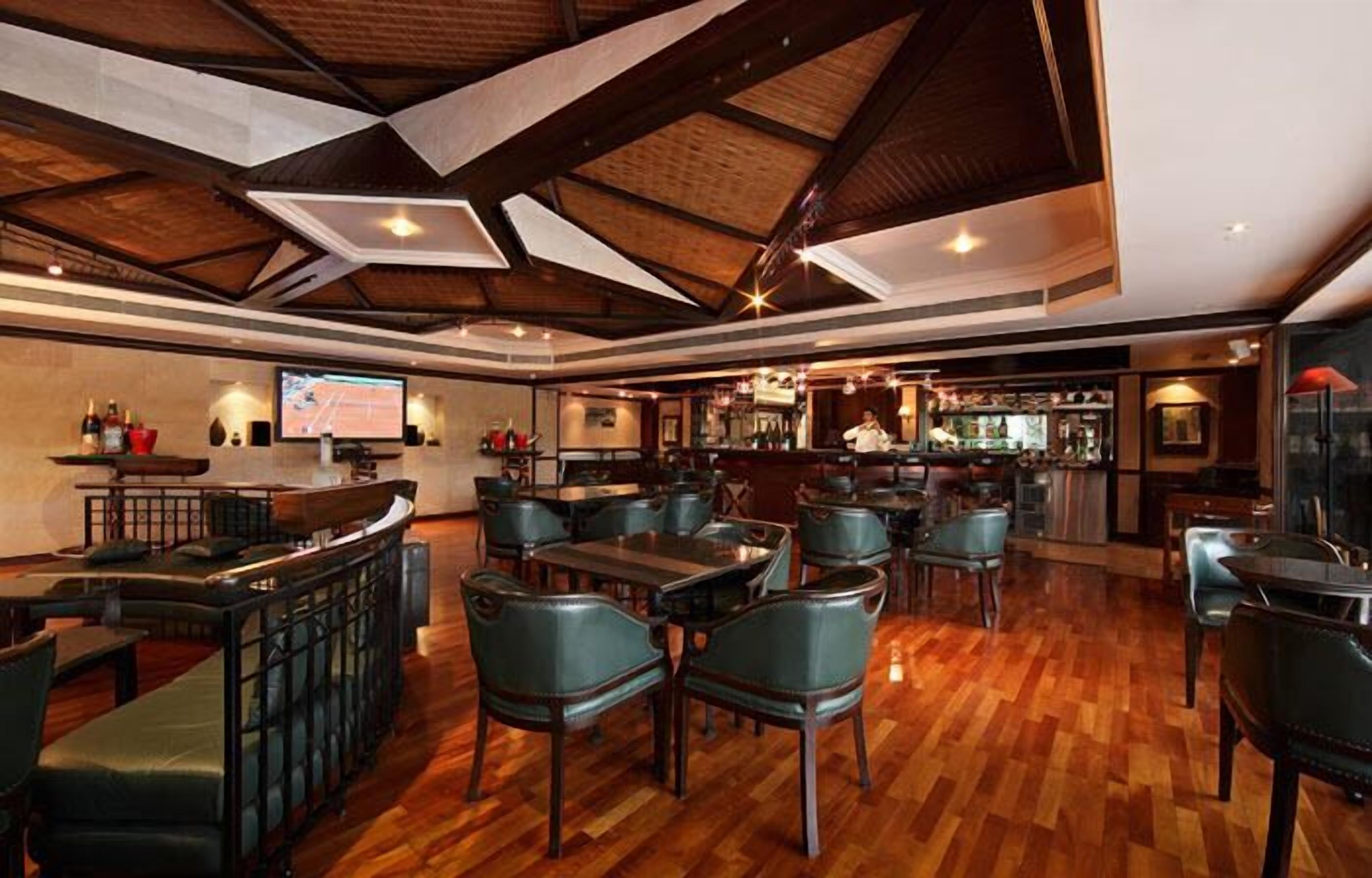 Un restaurant amb terres de fusta, cadires tapissades de verd i una zona de bar amb pantalla de televisió.