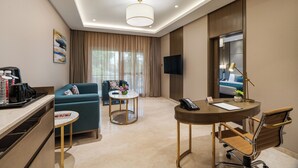 A hotel room with a sofa, a small round table, a desk with a chair, a TV, and a ceiling light. - Palm Meadows Resort (Bengaluru)