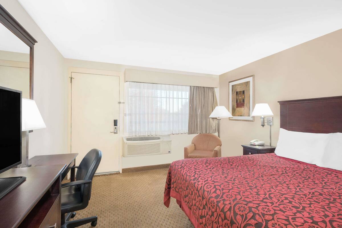A hotel room with a bed, a desk with a computer, a chair, a window with curtains, a painting on the wall, and a nightstand with a lamp.