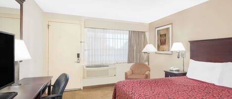 Habitación de hotel con cama, escritorio con computadora, silla, ventana con cortinas, un cuadro en la pared y mesita de noche con lámpara.