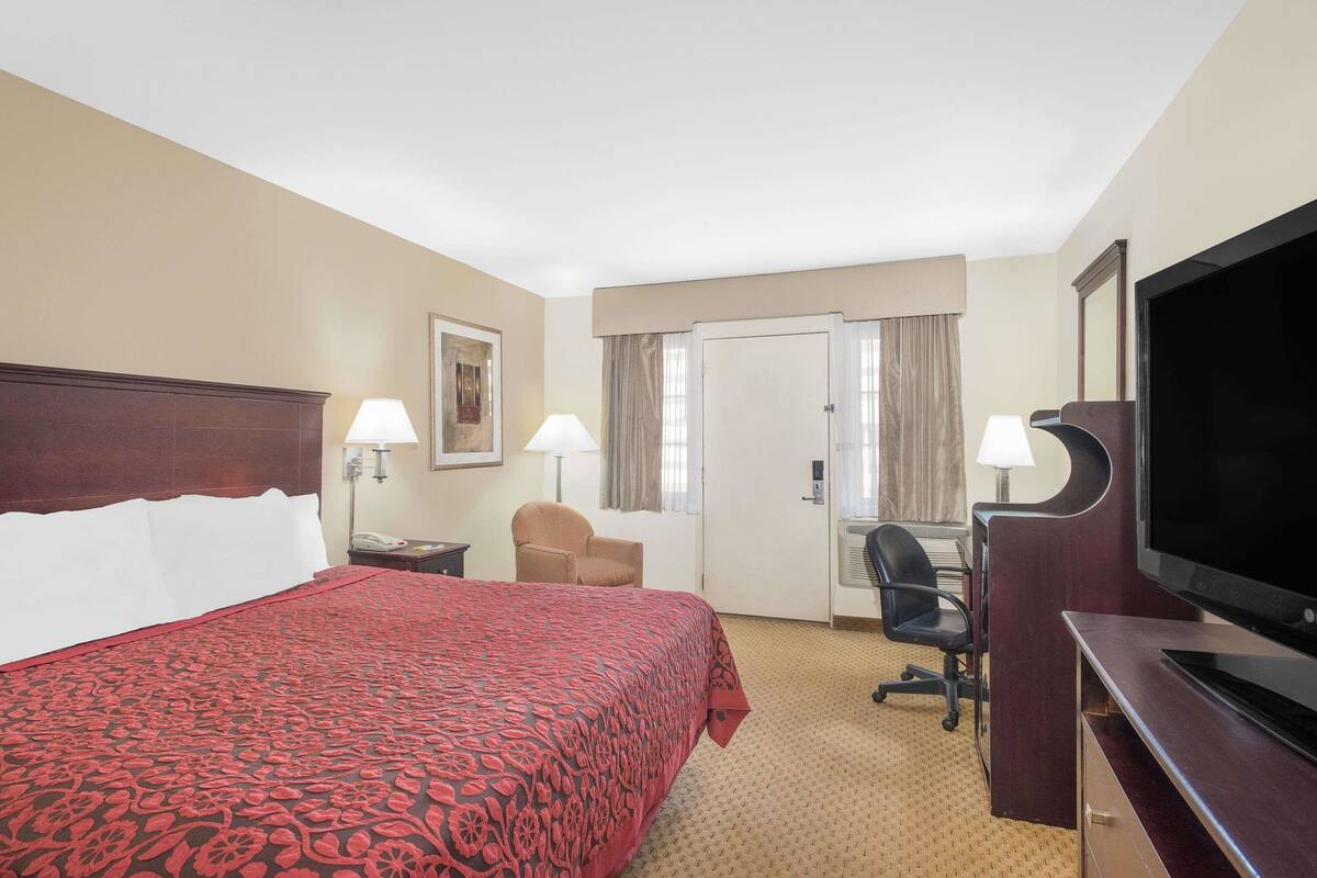 A hotel room with a large bed, a desk with a television, a chair, and a desk lamp.