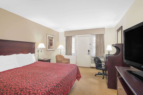 Quarto de hotel com uma cama grande, uma escrivaninha com televisão, uma cadeira e uma luminária de mesa.