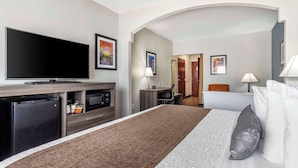 A hotel room with a large flat-screen TV, a refrigerator, a desk with a chair, a bed with a brown throw blanket, and a nightstand with a lamp. - Best Western Plus Victoria Inn & Suites (Victoria)