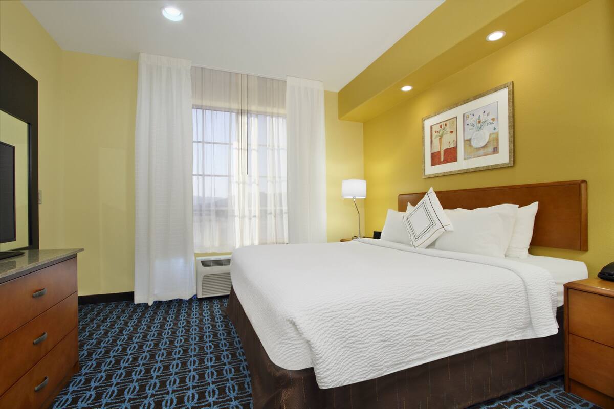 Ein Hotelzimmer mit einem großen Bett, einem Schreibtisch, einem Sessel, einem Fernseher und einem Fenster mit Vorhängen.