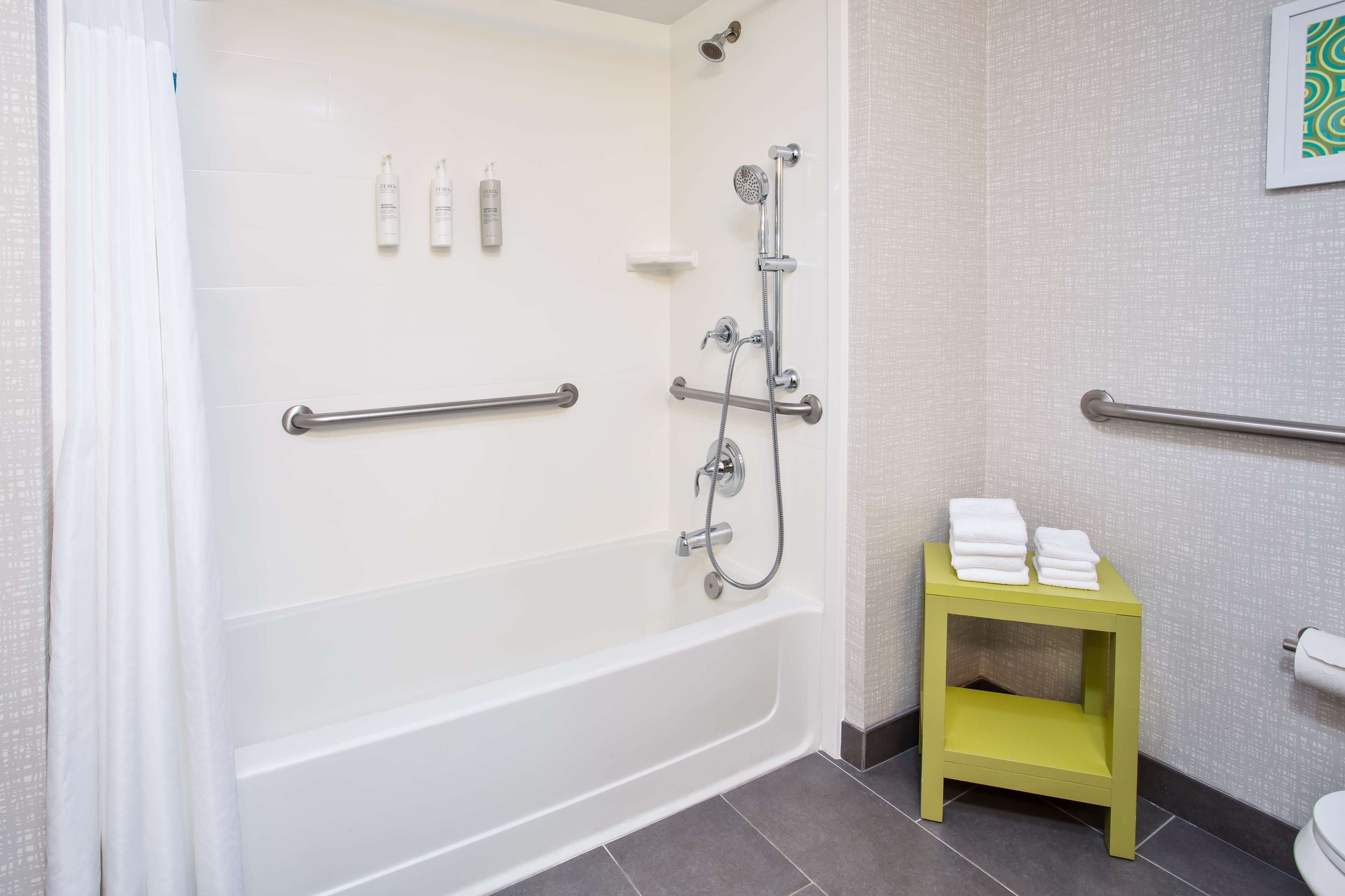Une salle de bain avec une baignoire blanche, des barres d’appui, une pomme de douche et une petite étagère jaune sur laquelle sont posées des serviettes.