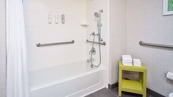 A bathroom with a white bathtub, grab bars, a showerhead, and a small yellow shelf with towels.