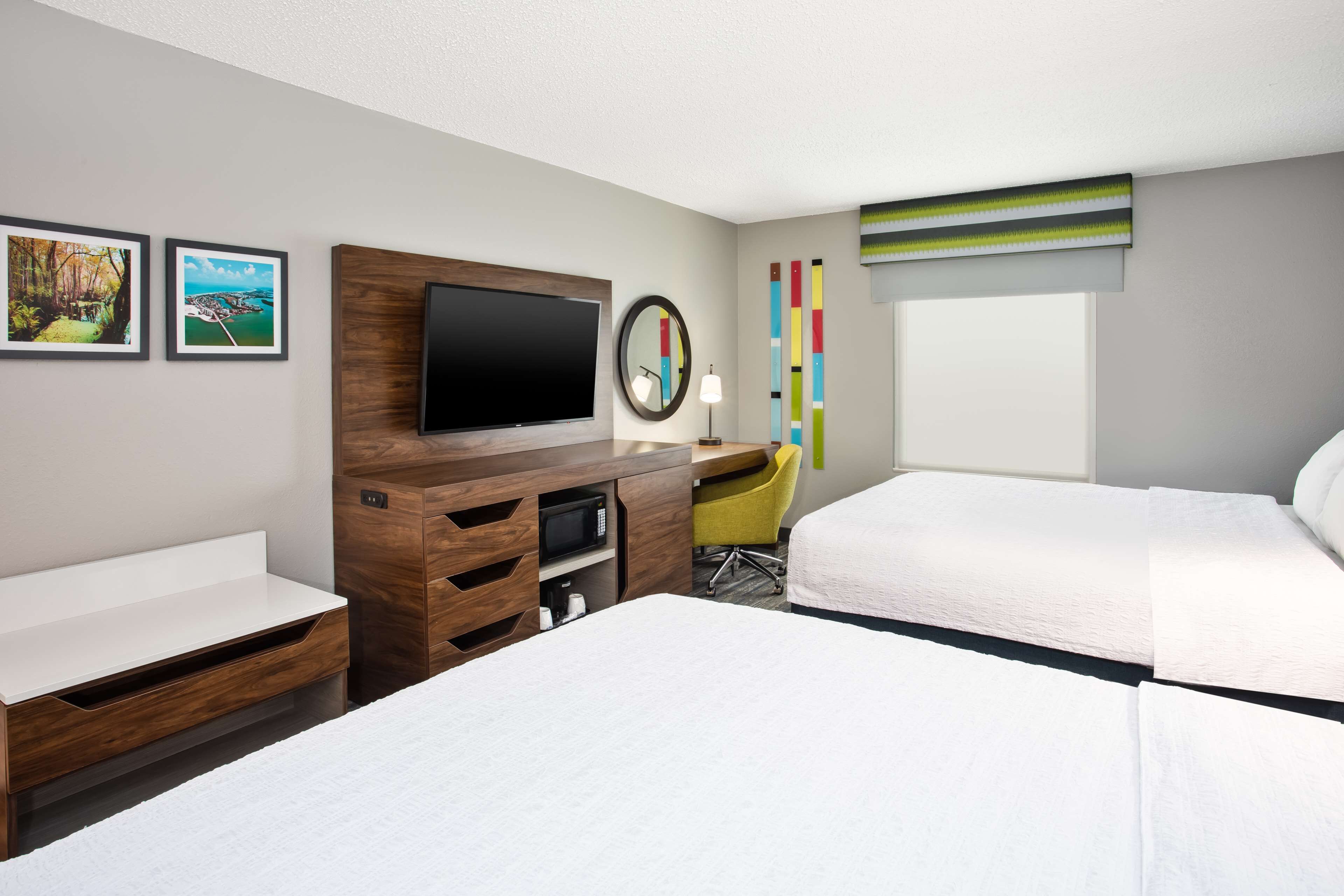A hotel room with two beds, a desk, a flat-screen TV, a mirror, and a window with blinds.