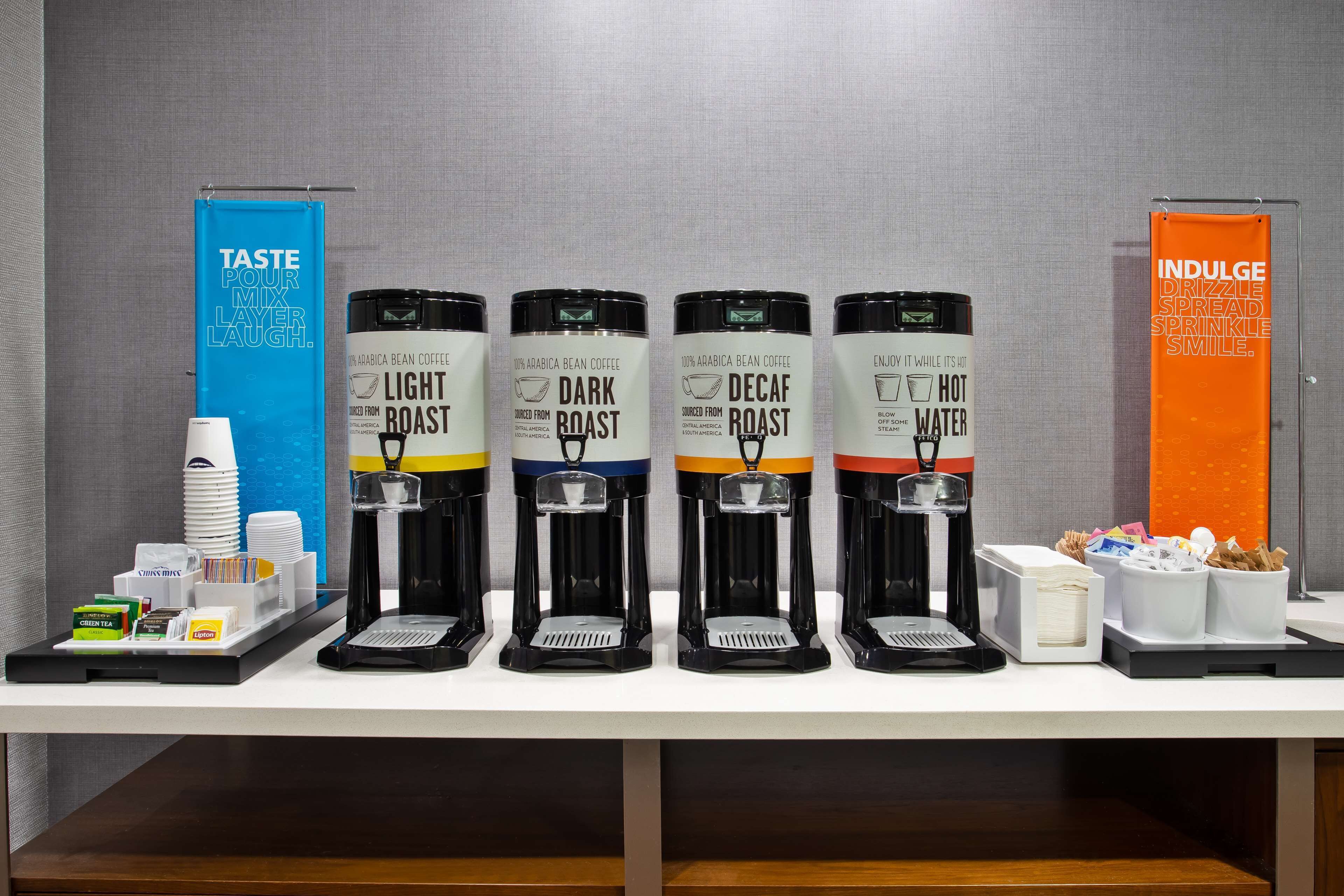A coffee station with four coffee dispensers labeled 'Light Roast,' 'Dark Roast,' 'Decaf Roast,' and 'Hot Water.' There are also coffee cups, a coffee maker, and a sign that reads 'Taste Pour Mix Layer Laugh.'