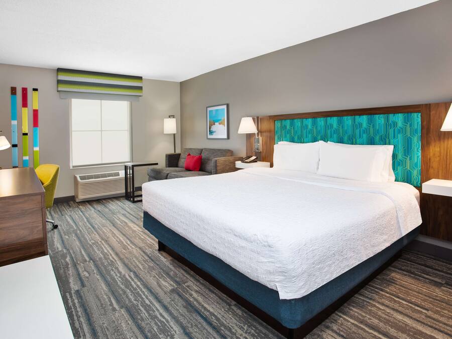 A hotel room with a large bed, a desk, a chair, a window, and a wall with colorful vertical stripes.