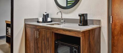 A bathroom with a wooden vanity, a sink, a coffee maker, a microwave, and a round mirror.