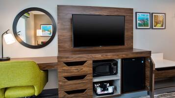 A modern hotel room with a wooden desk, a flat-screen TV, a microwave, and a small refrigerator. There is a green chair and a round mirror on the wall.
