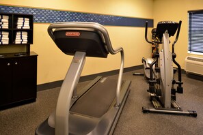 Un gimnasio con cinta de correr, elíptica y pesas. Hay una pared con diseño hexagonal y un estante con toallas.