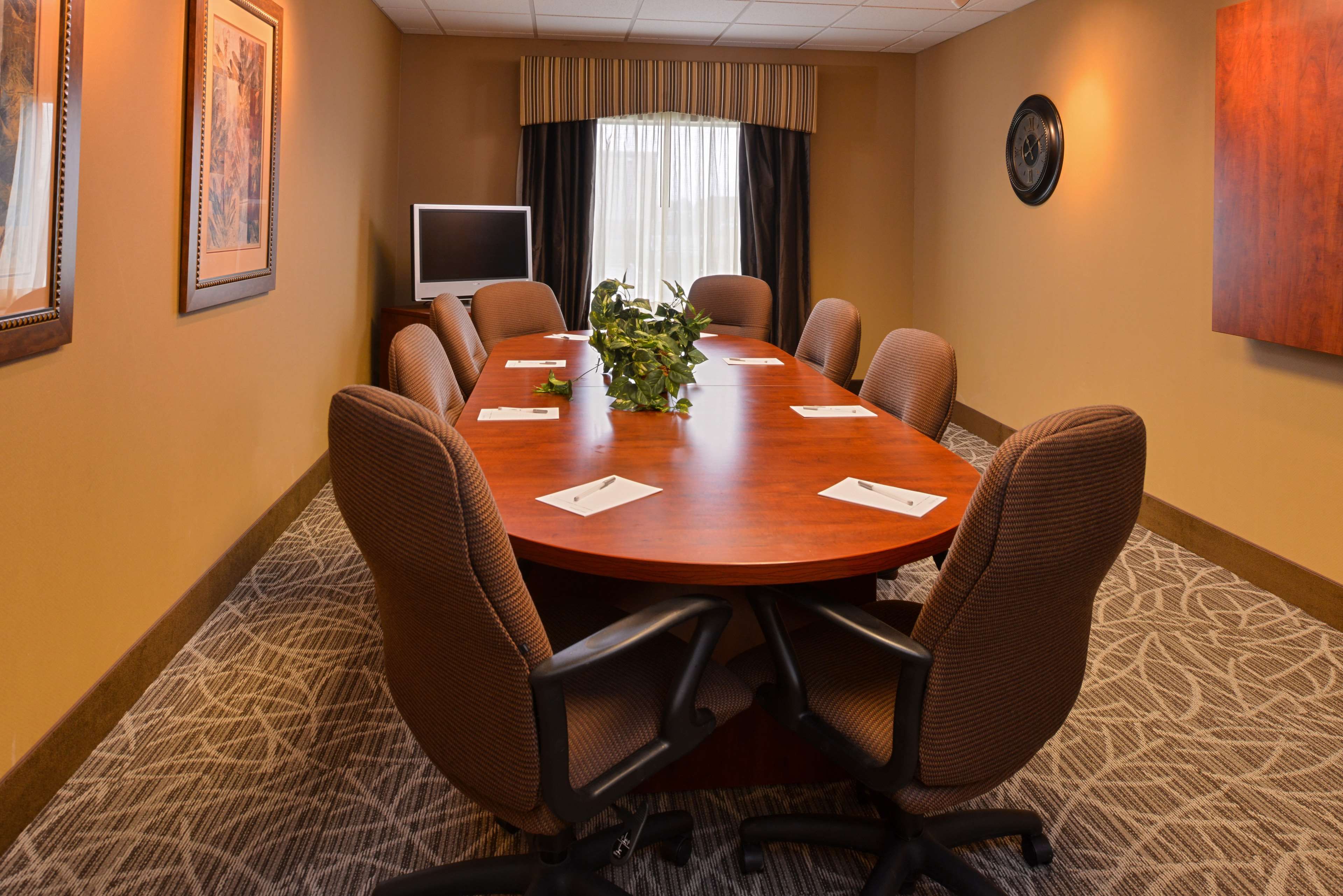 A conference room with a long table, six chairs, a plant, a clock, and a television on a stand.