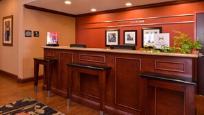 A reception desk with a 'Welcome to Litchfield' sign, framed pictures, a small plant, and a notice about a small location. - Hampton Inn by Hilton Litchfield (Litchfield)