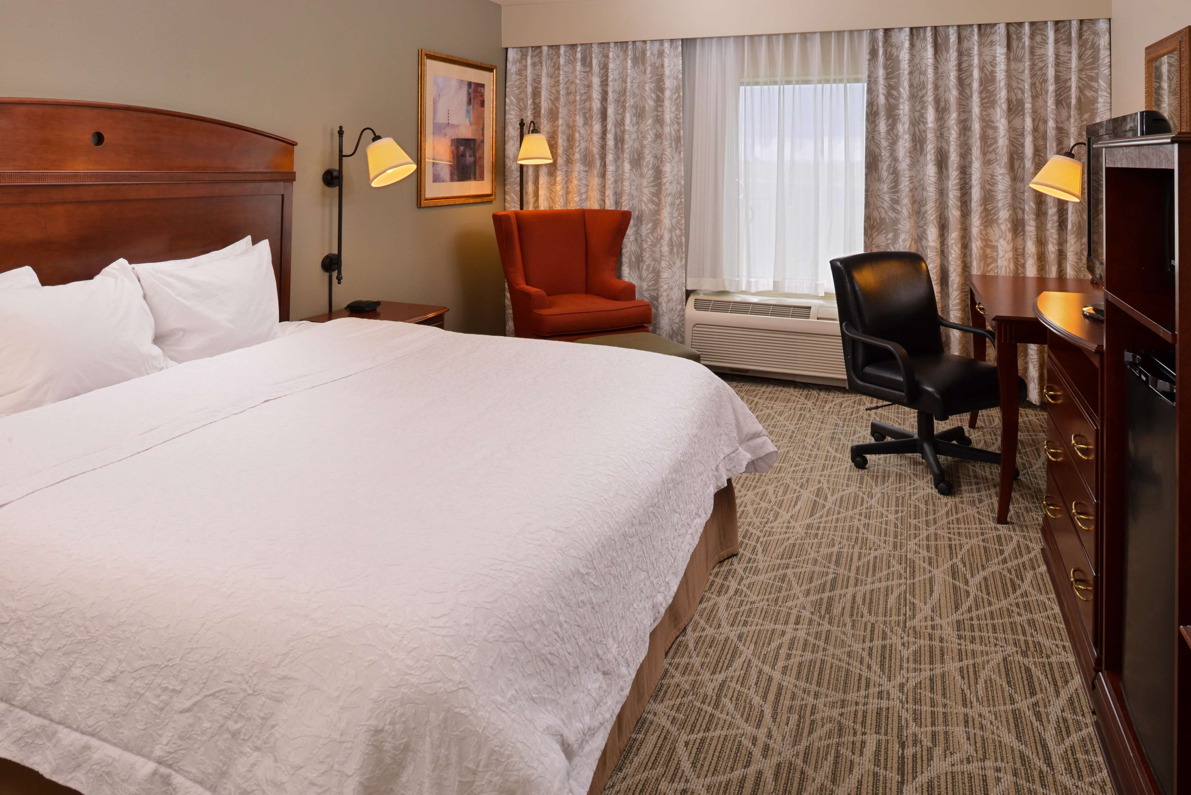 A hotel room with a large bed, two chairs, a desk with a computer, and a window with curtains.