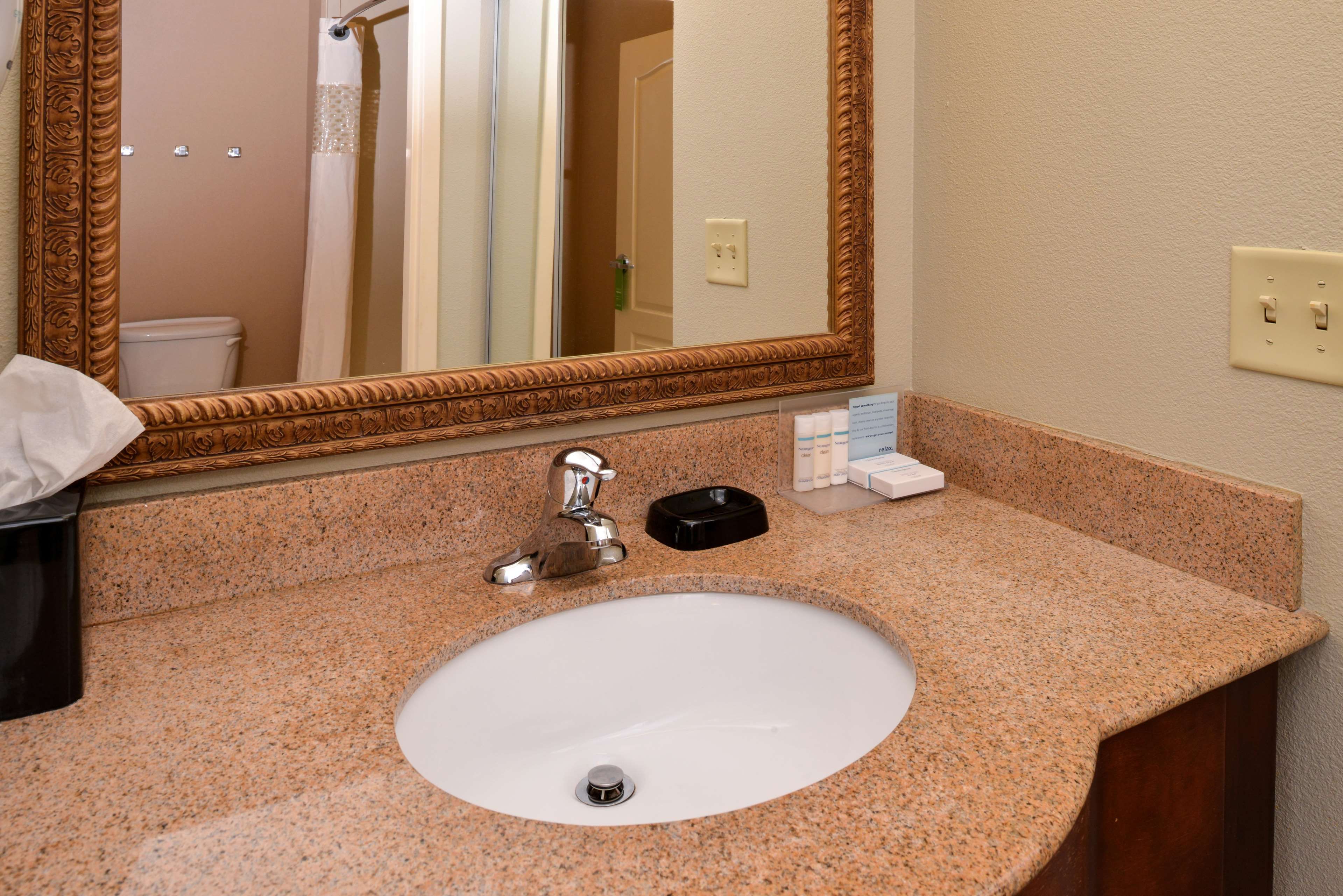 A bathroom with a granite countertop, a white sink, a mirror with an ornate frame, and toiletries on the counter.