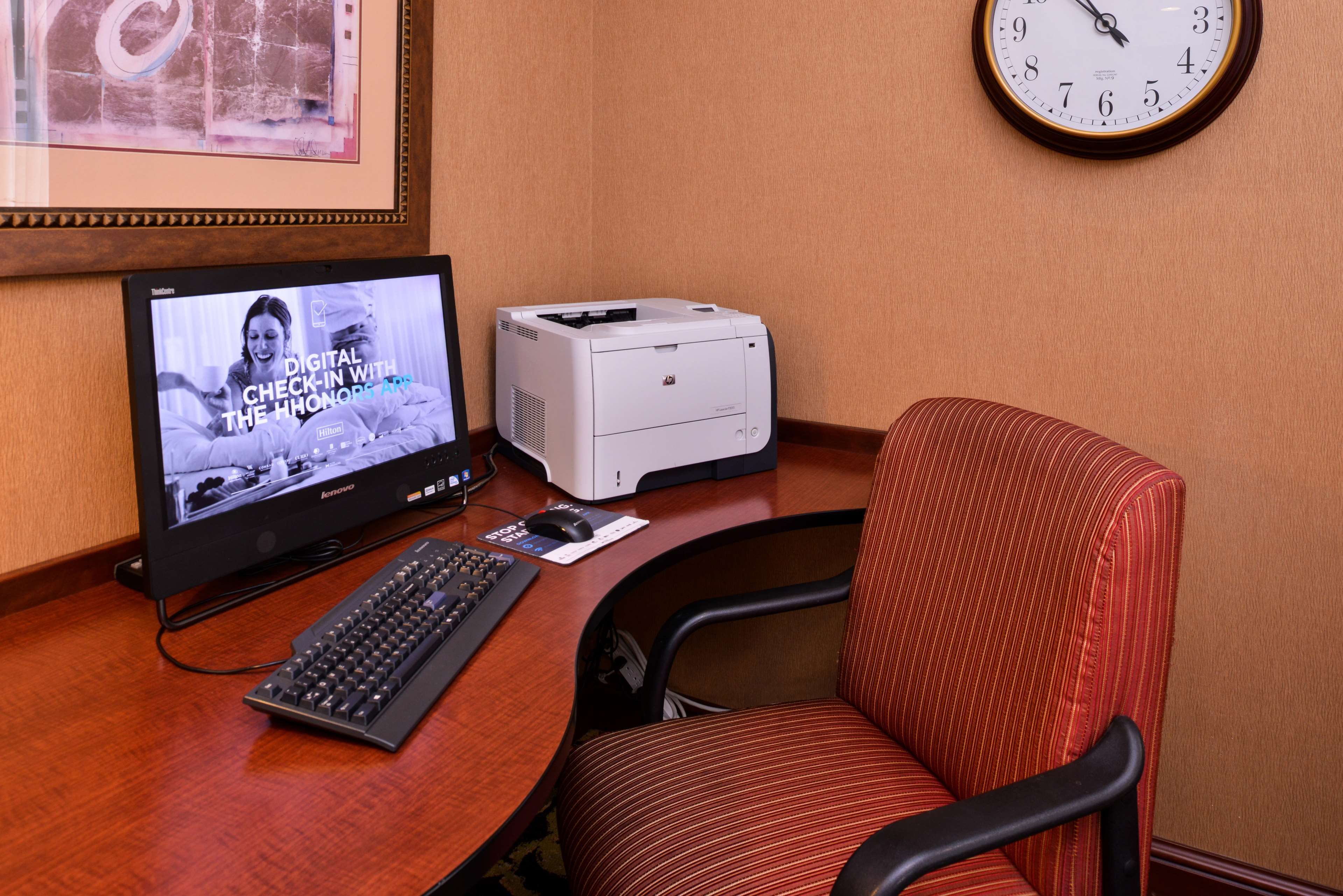 Habitación de hotel con escritorio, computadora de escritorio, impresora, reloj y un cuadro en la pared.