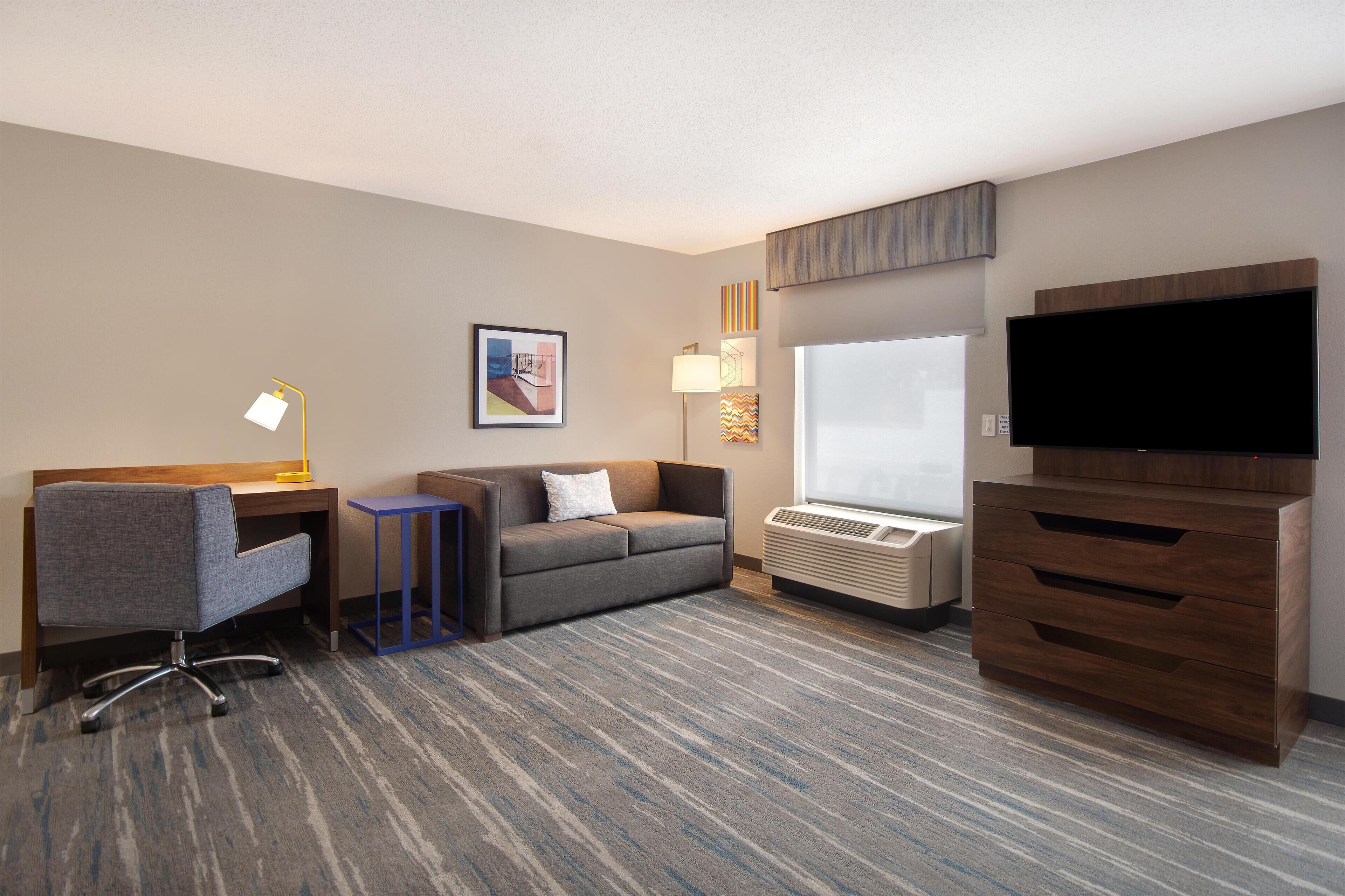 A hotel room with a desk, a sofa, a TV, and a dresser.