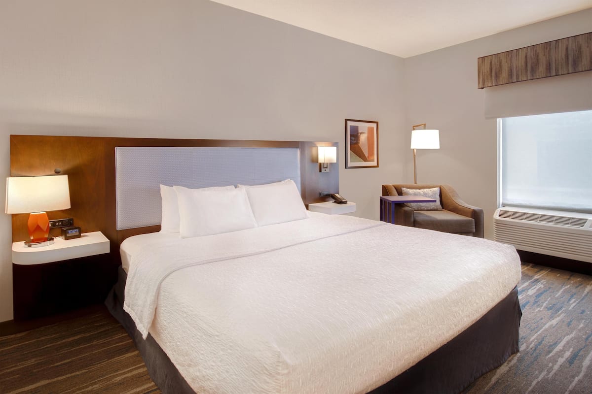Una habitación de hotel con una cama grande, una mesita de noche con lámpara, una silla, un cuadro en la pared y una ventana con aire acondicionado.