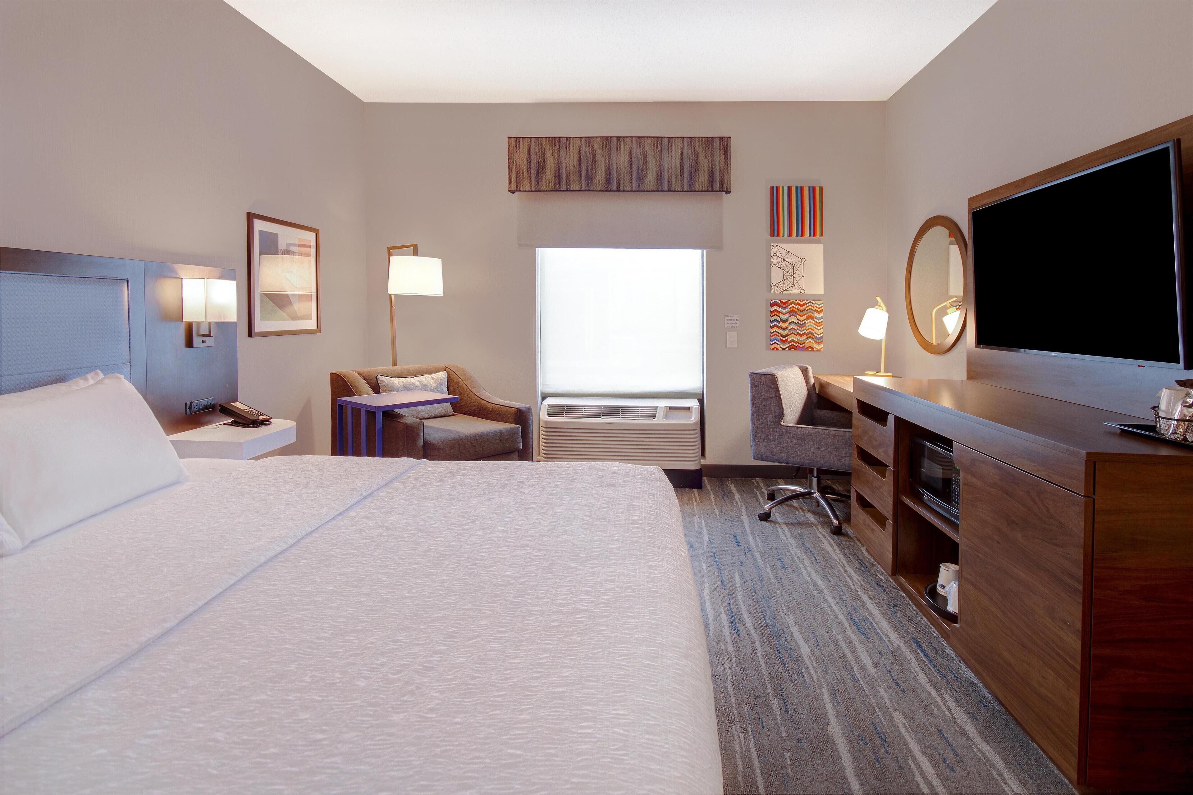 Quarto de hotel com uma cama grande, uma escrivaninha, uma cadeira, uma televisão e um ar condicionado.