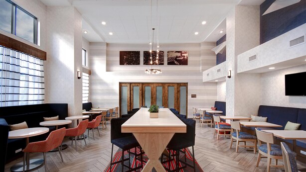 A modern dining area with a central wooden table, surrounded by chairs and booths, featuring a herringbone floor pattern and a red and black rug. - Hampton Inn & Suites Dayton - Vandalia (Dayton)