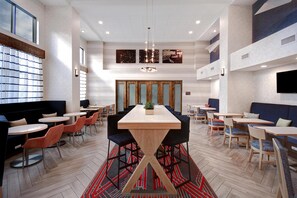 A modern dining area with a central wooden table, surrounded by chairs and booths, featuring a herringbone floor pattern and a red and black rug. - Hampton Inn & Suites Dayton - Vandalia (Dayton)