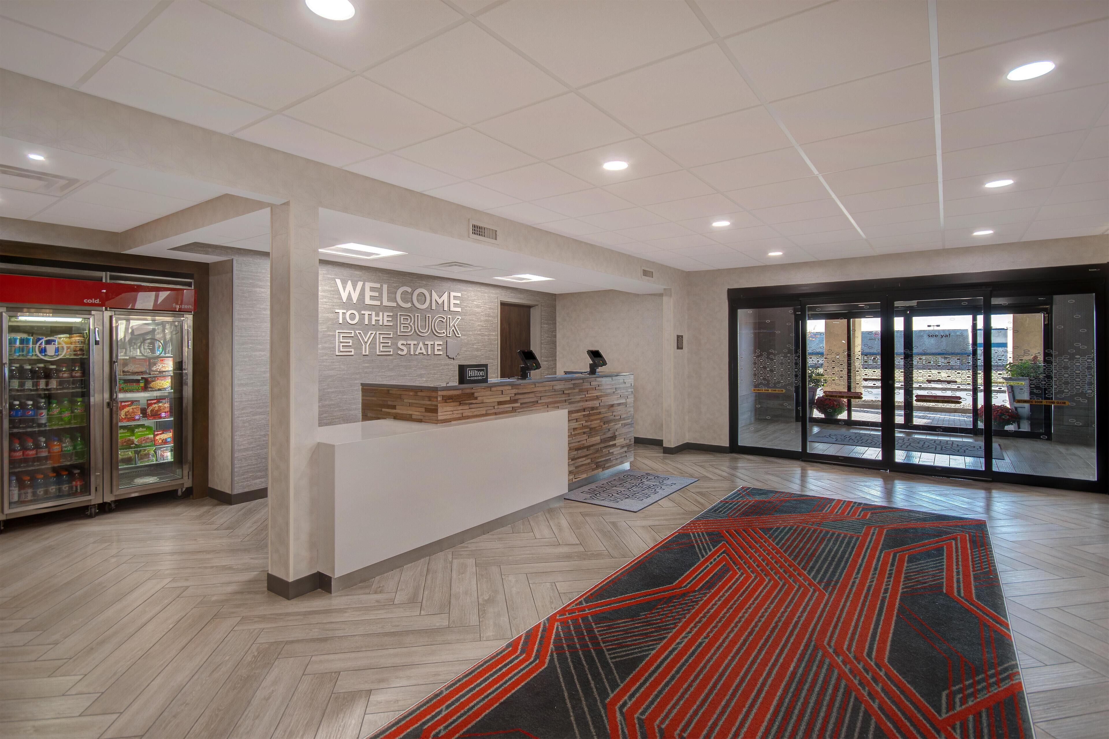 A reception area with a welcome sign, a counter, a refrigerator, and a patterned carpet.