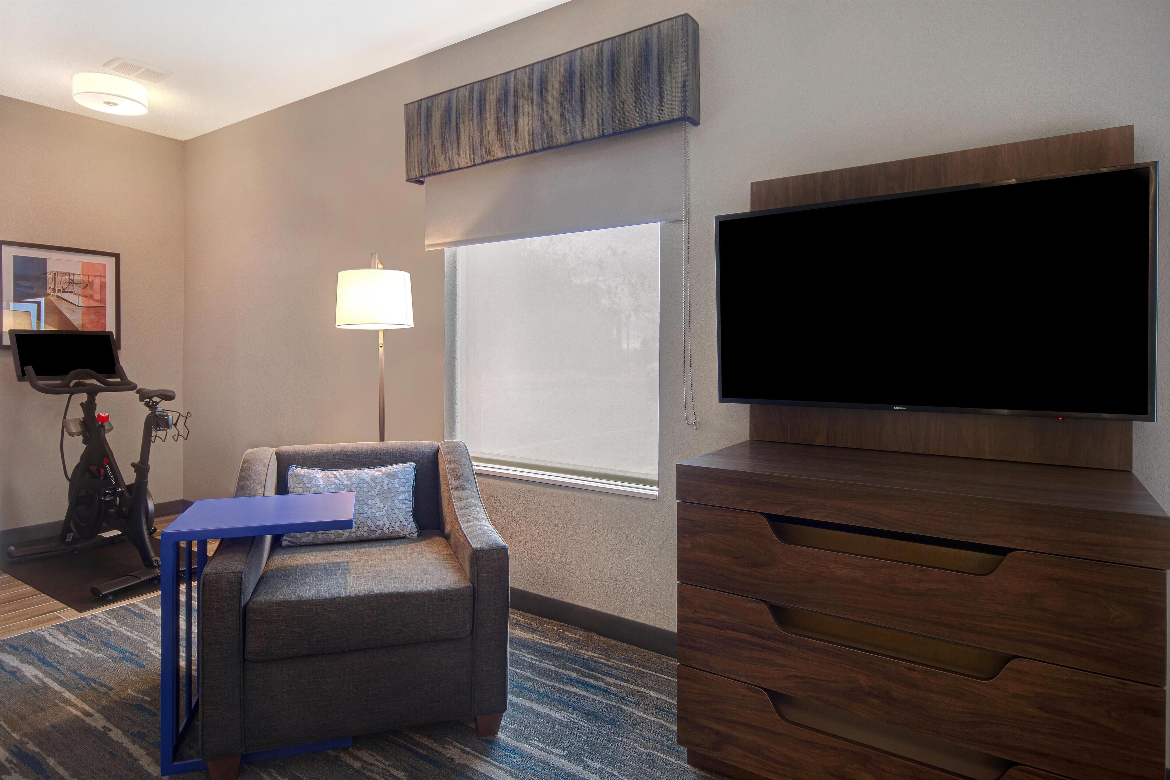 A hotel room with a gray sofa, a flat-screen TV, a wooden dresser, a lamp, and a treadmill.