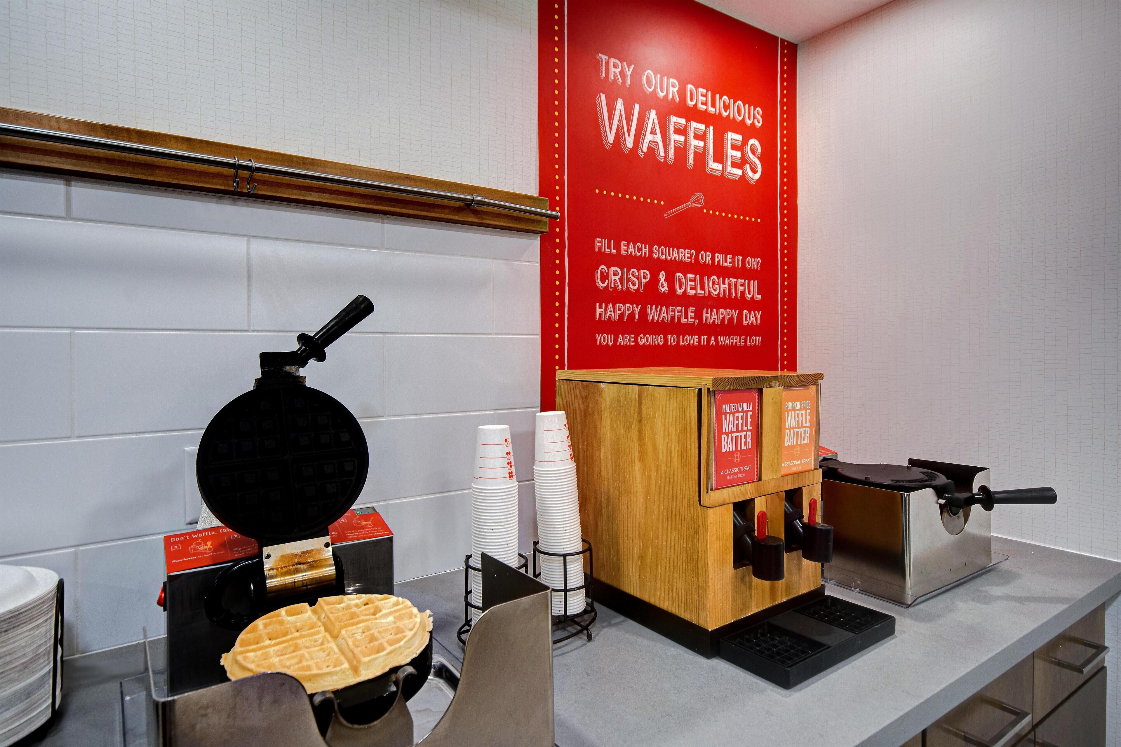 A waffle station with a waffle maker, stack of waffle plates, and a waffle batter dispenser.