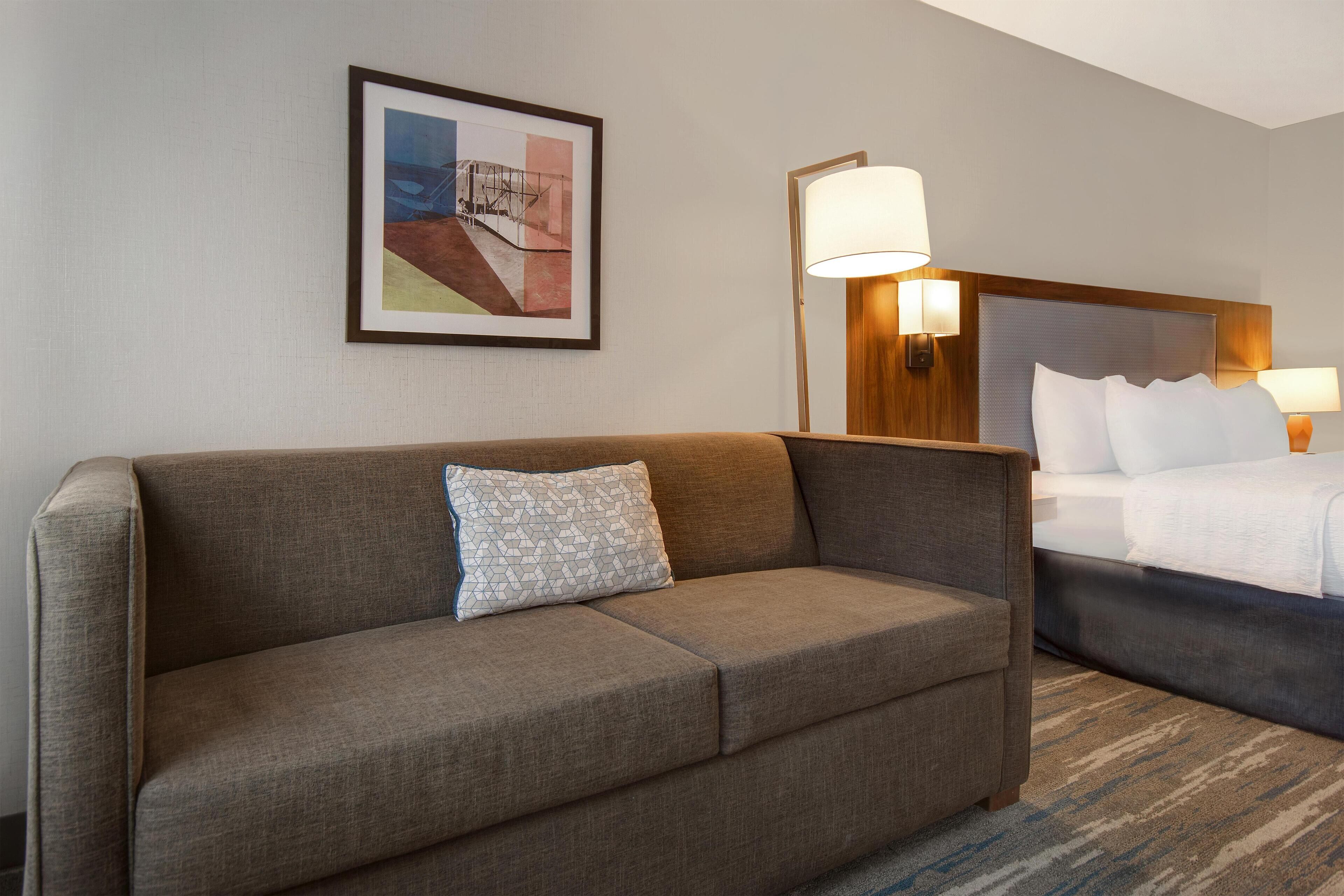Une chambre d’hôtel avec un canapé, un lit, un tableau encadré au mur et deux lampes fixées au mur.