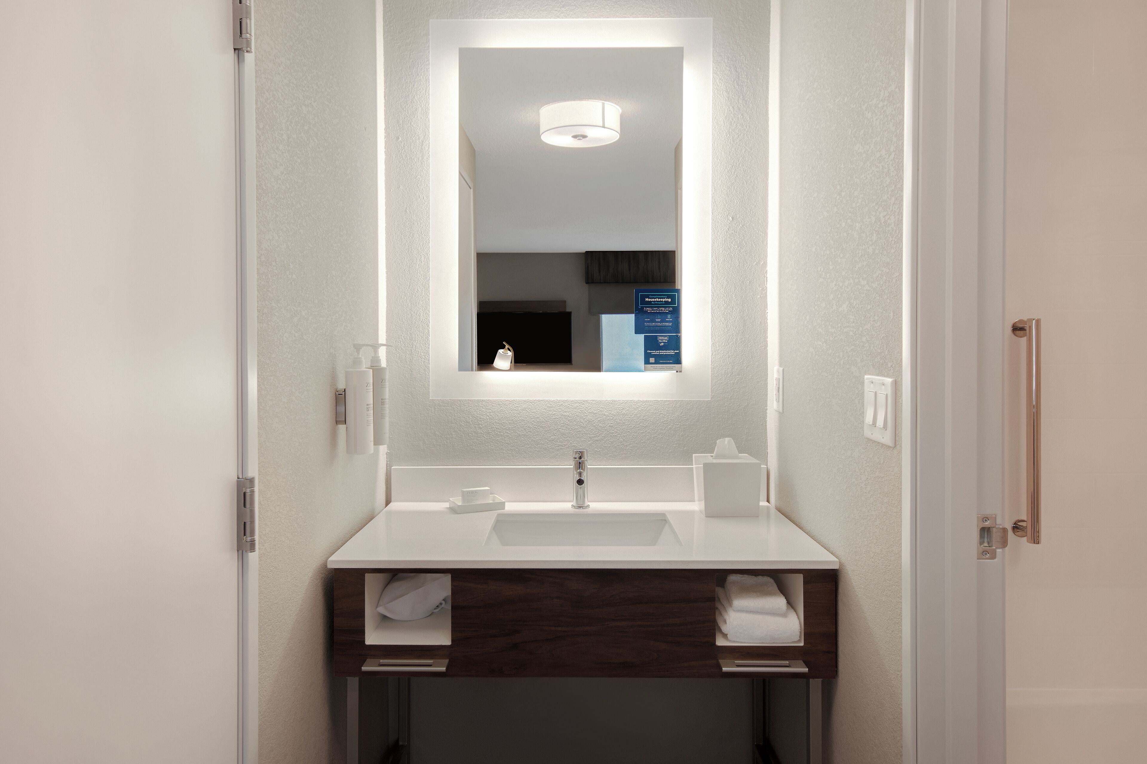Een badkamer met een witte wastafel, een donker houten wastafelmeubel en een spiegel met ingebouwde verlichting.