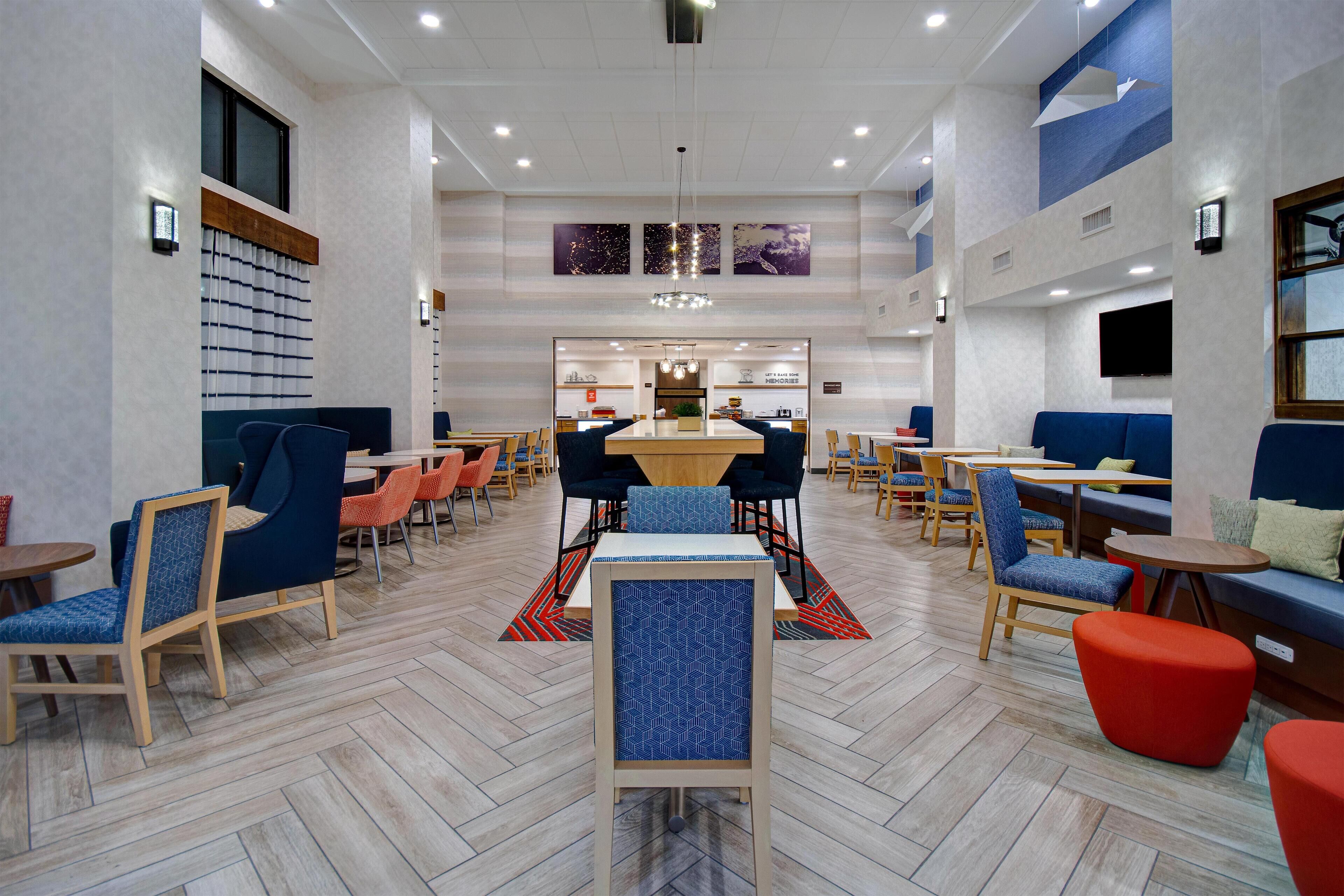 A modern hotel lobby with a central dining area, multiple seating options, and a large TV screen on the wall.