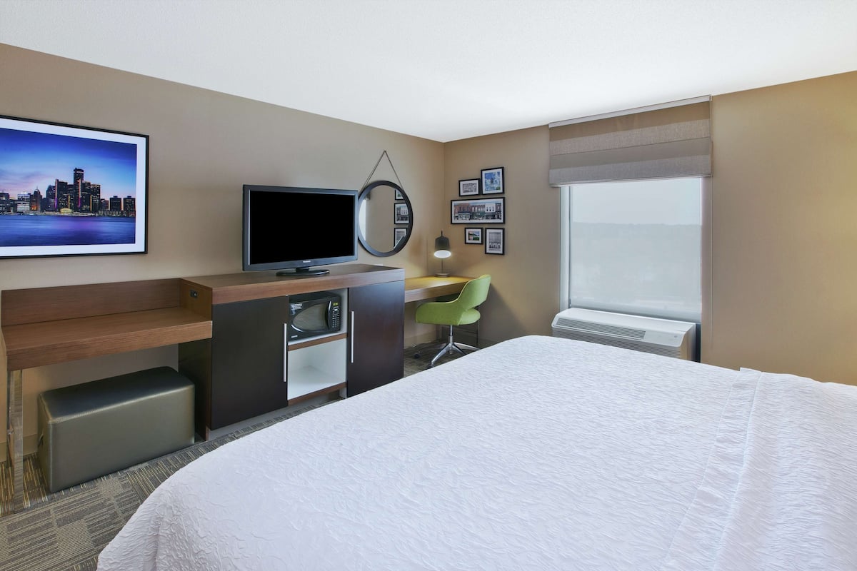 A hotel room with a bed, a desk with a chair, a television, a microwave, a mirror, framed pictures on the wall, and a window.