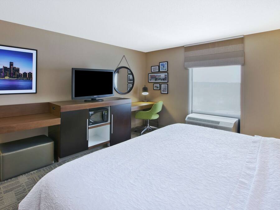 Una habitación de hotel con una cama, un escritorio con una silla, un televisor, un microondas, un espejo, cuadros enmarcados en la pared y una ventana.