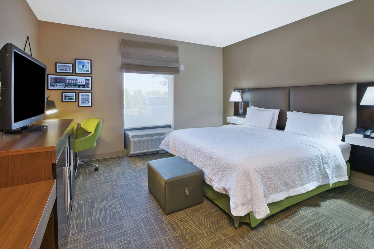 A hotel room with a bed, a desk with a television, a chair, and a window with a view of the outside.