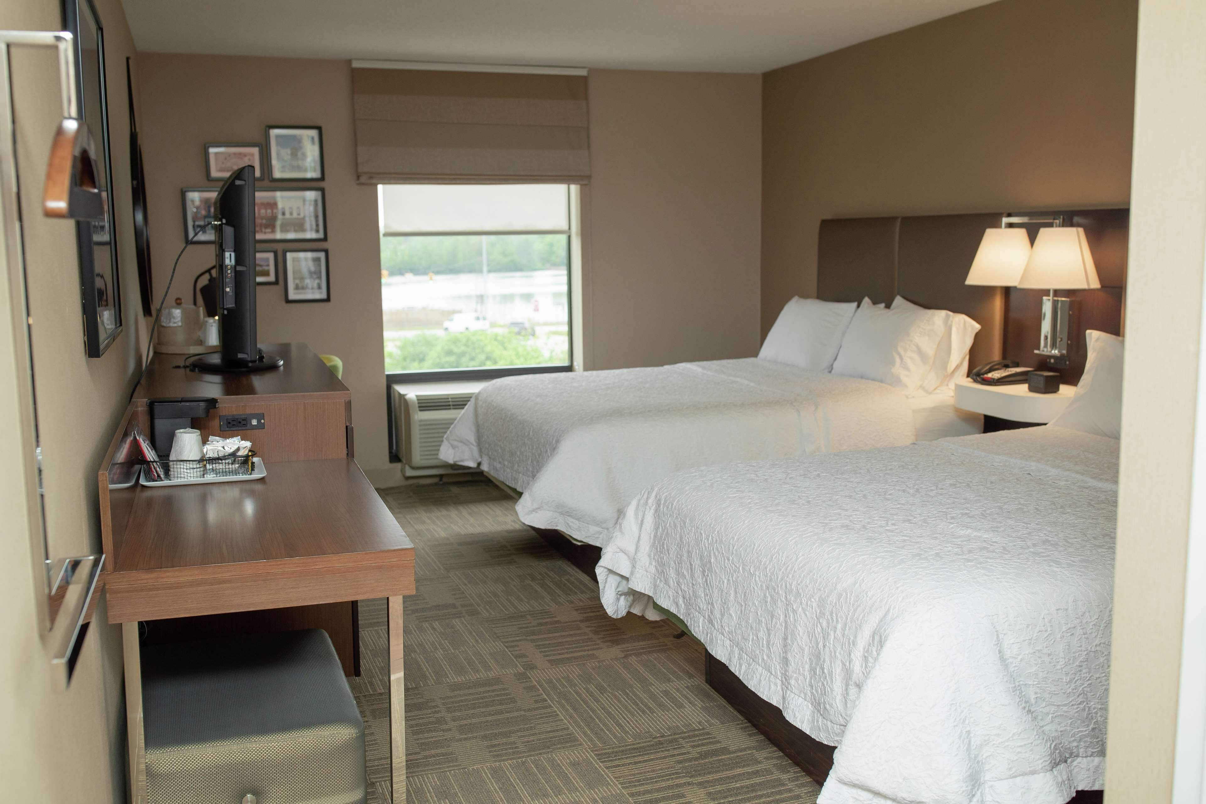 A hotel room with two beds, a desk, a chair, a TV, and a window with a view of the outside.