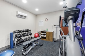 Centro de fitness com esteira, banco de musculação, halteres, uma bola de exercício vermelha e ar condicionado fixado na parede.