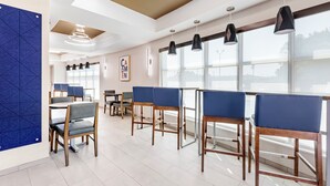 A modern dining area with high tables and blue cushioned chairs, large windows, and contemporary lighting fixtures. - Holiday Inn Express Hotel & Suites Camden-I20 (Hwy 521) by IHG (Camden)