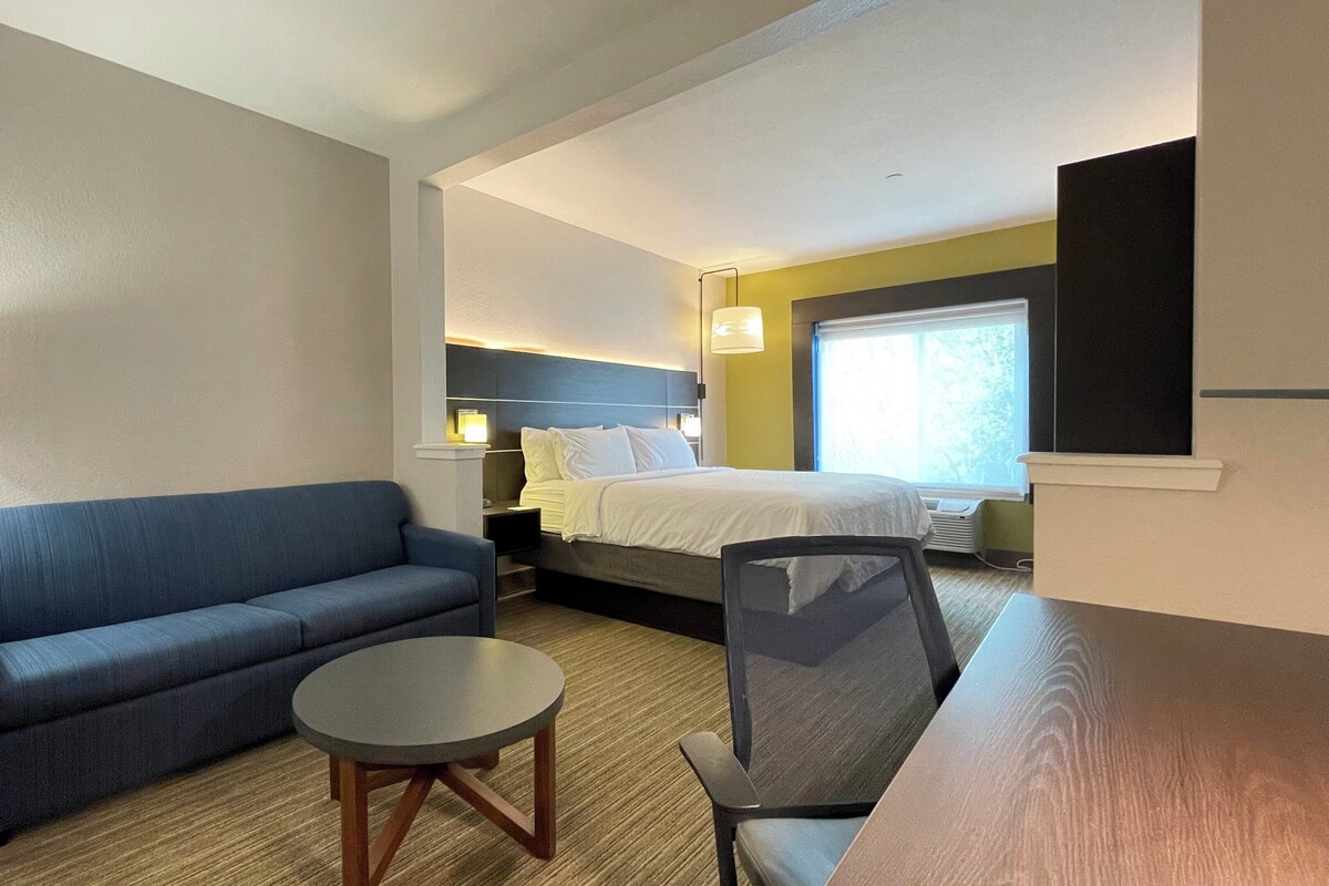 A hotel room with a blue sofa, a round table, a bed with white bedding, and a nightstand with a lamp.