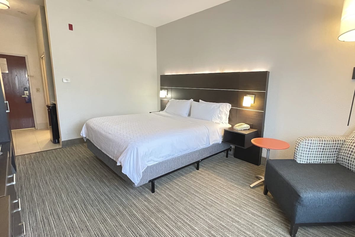 A hotel room with a bed, a bedside table with a lamp, a chair, and a small table.