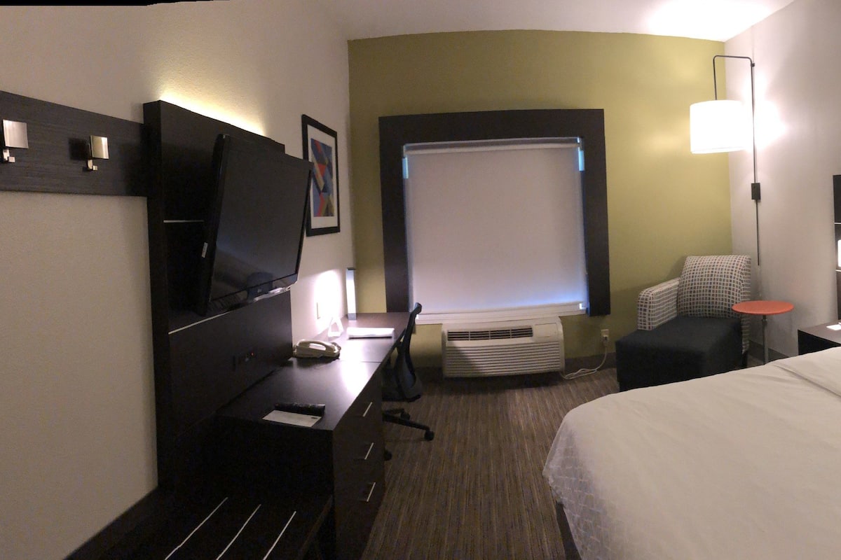 A hotel room with a bed, a desk, a chair, a TV, a bedside table, and a lamp.